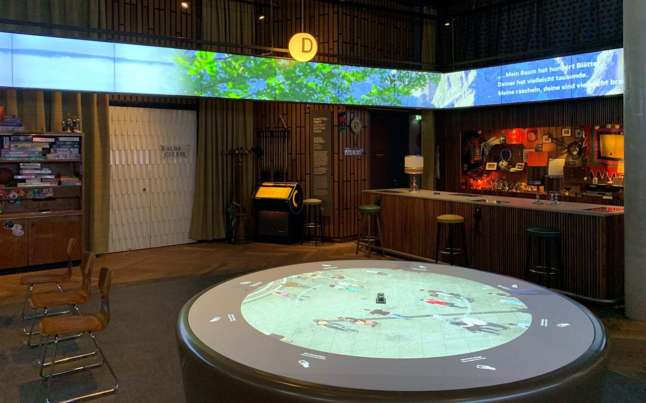 Runder interaktiver Tisch mit Projektion in einem Ausstellungsraum, darüber ein umlaufendes Band aus Bildschirmen