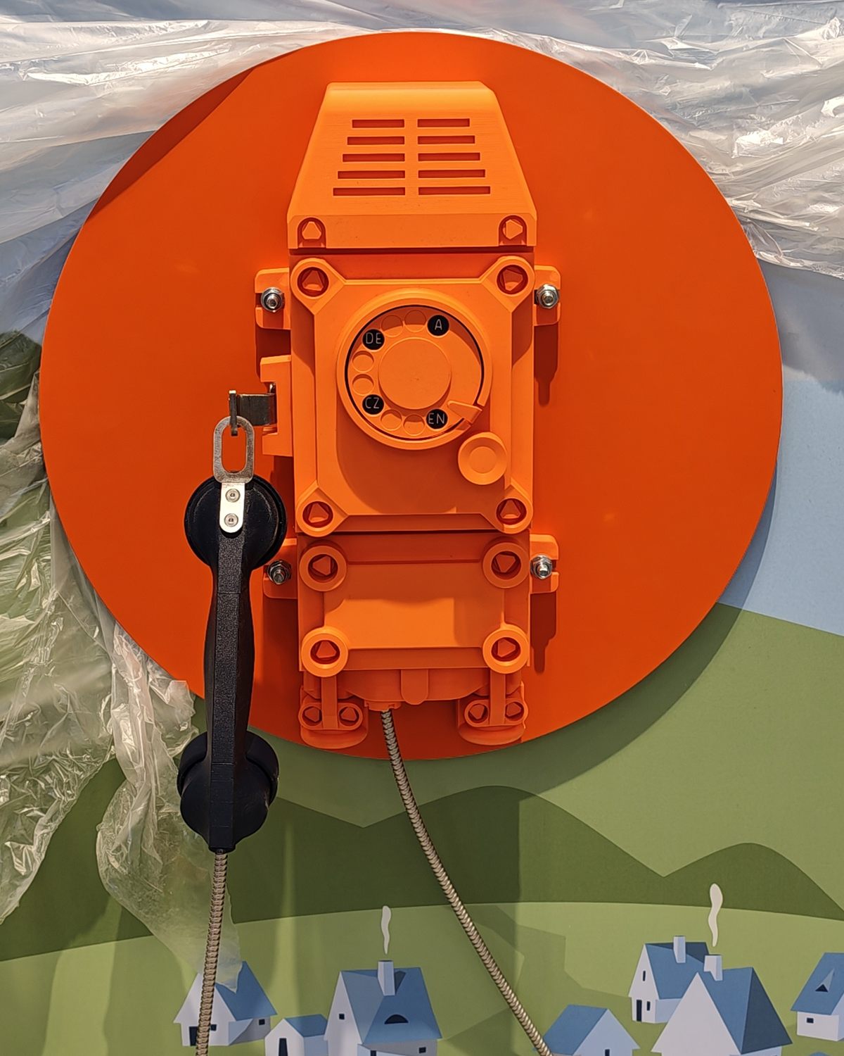Nahaufnahme des gedruckten Grubentelefons: Gehäuse mit Lüftungsschlitzen und Schraubenköpfen, Wählscheibe mit den Kürzeln DE, CZ und EN, daneben der Hörer am Haken und ein Metallschlauch als Kabel