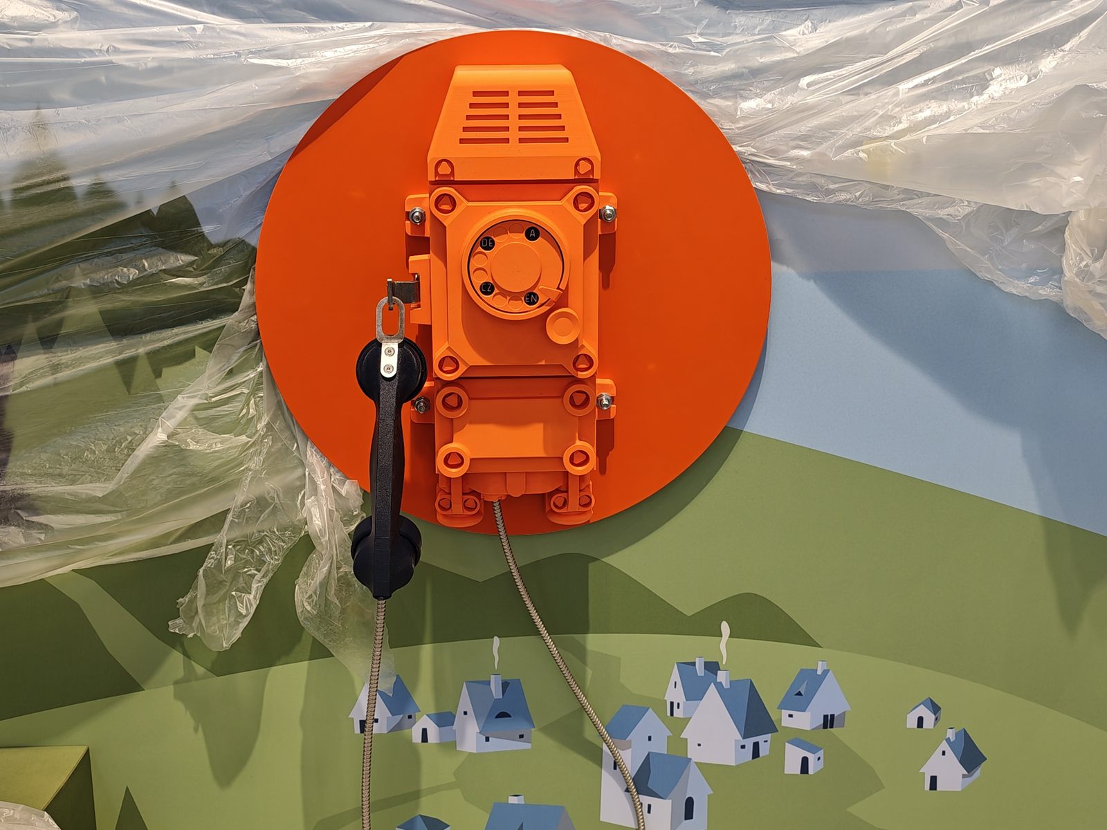 Orangefarbenes, 3D-gedrucktes Grubentelefon mit Wählscheibe und schwarzem Hörer auf einer runden Platte an einer illustrierten Ausstellungswand mit Hügeln und Dorf; ringsum noch Schutzfolie vom Aufbau