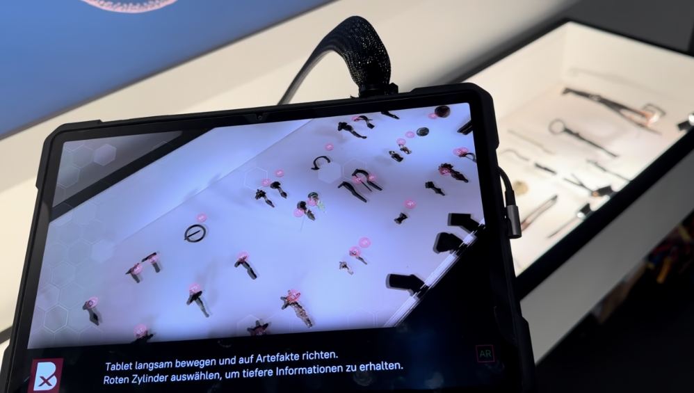 Ein Tablet zeigt die Vitrine mit Grabfunden; über den Objekten schweben virtuelle Knöpfe