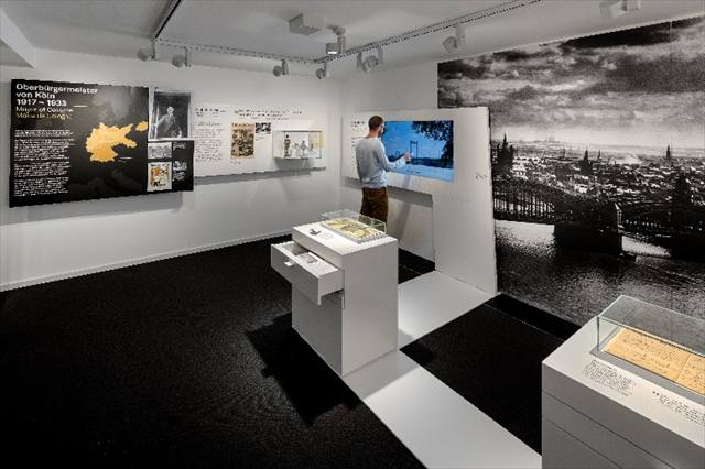Ausstellungsraum mit Kölner Stadtpanorama, Vitrinen und Besucher an einer Wand-Medienstation