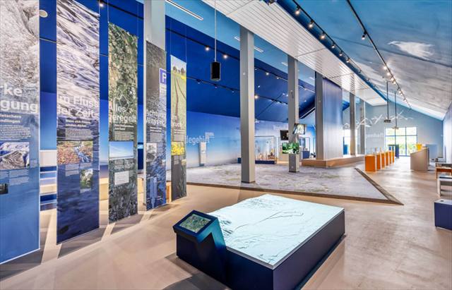Ausstellungshalle mit Landschaftsbannern, im Vordergrund ein Reliefmodell mit Touchscreen