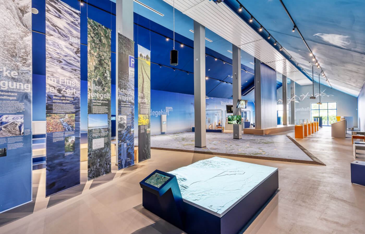 Ausstellungshalle mit Landschaftsbannern, im Vordergrund ein Reliefmodell mit Touchscreen