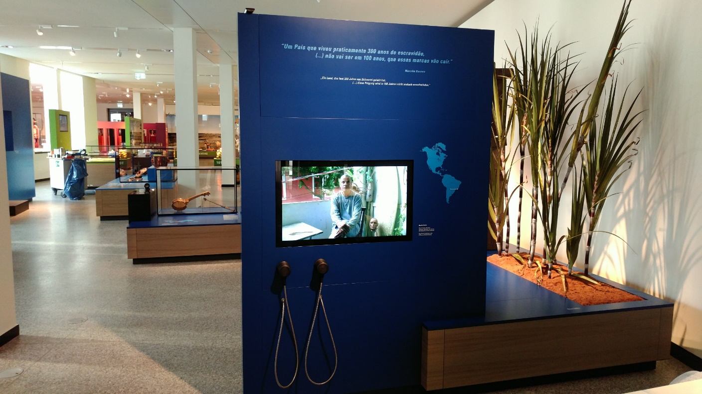 Blaue Ausstellungswand mit Videostation, zwei Kopfhörern und Pflanzen in der Ausstellung „Amerika“