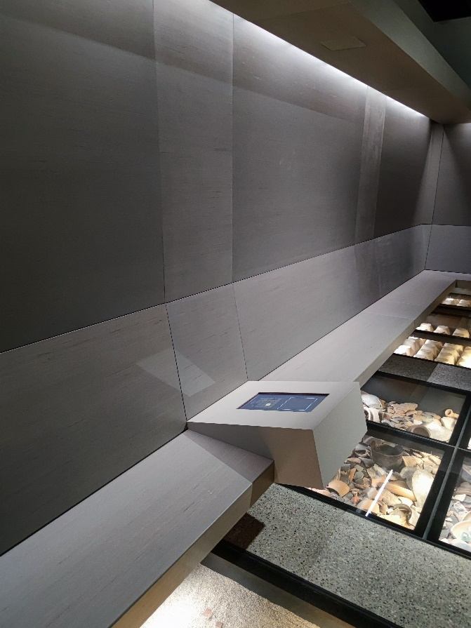 Ausstellungsnische mit Touchscreen-Pult an einer Sitzbank und Bodenvitrinen mit Funden