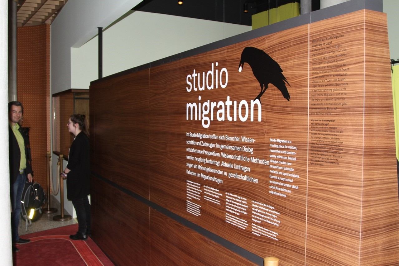 Eingangswand des „Studio Migration“ mit Holzverkleidung, Schriftzug und Rabenmotiv