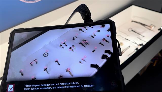 Tablet mit AR-App vor einer Vitrine, eingeblendete Markierungen über den Grabfunden
