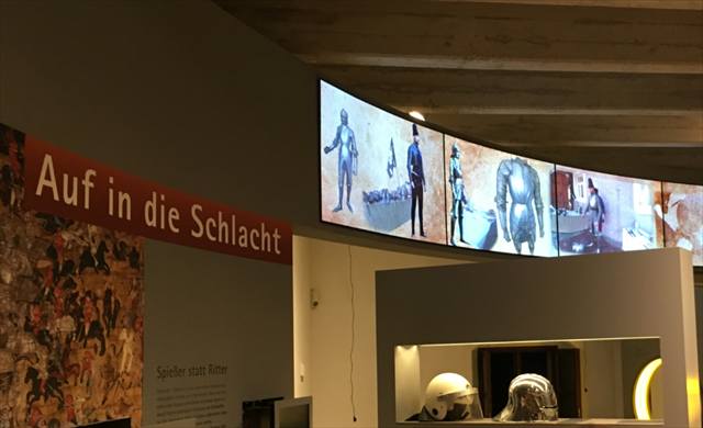 Ausstellungsbereich „Auf in die Schlacht“ mit gebogener Bildschirmreihe und Rüstungsexponaten
