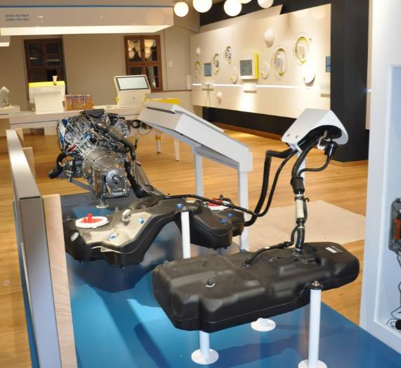 Ausstellungsraum mit Fahrzeugmotor und Tanksystemen als Exponat, im Hintergrund weitere Stationen