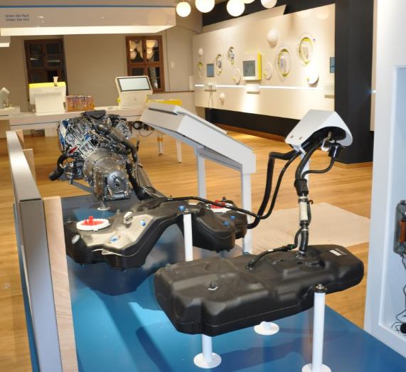 Ausstellungsraum mit Fahrzeugmotor und Tanksystemen als Exponat, im Hintergrund weitere Stationen