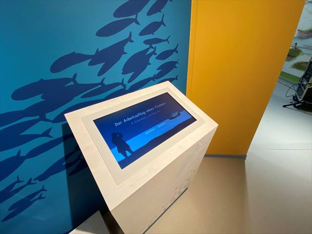 Touchscreen-Pult vor blauer Wand mit Fischschwarm-Grafik