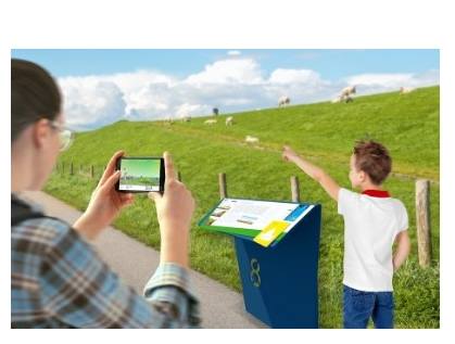 Person hält am Deich ein Smartphone mit App-Ansicht, daneben eine Infostele und ein Kind