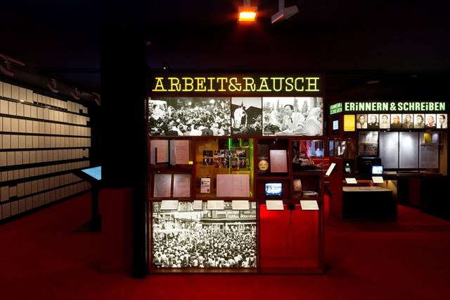 Themeninsel „Arbeit & Rausch“ mit Fotos, Vitrinen und kleinen Monitoren auf rotem Boden