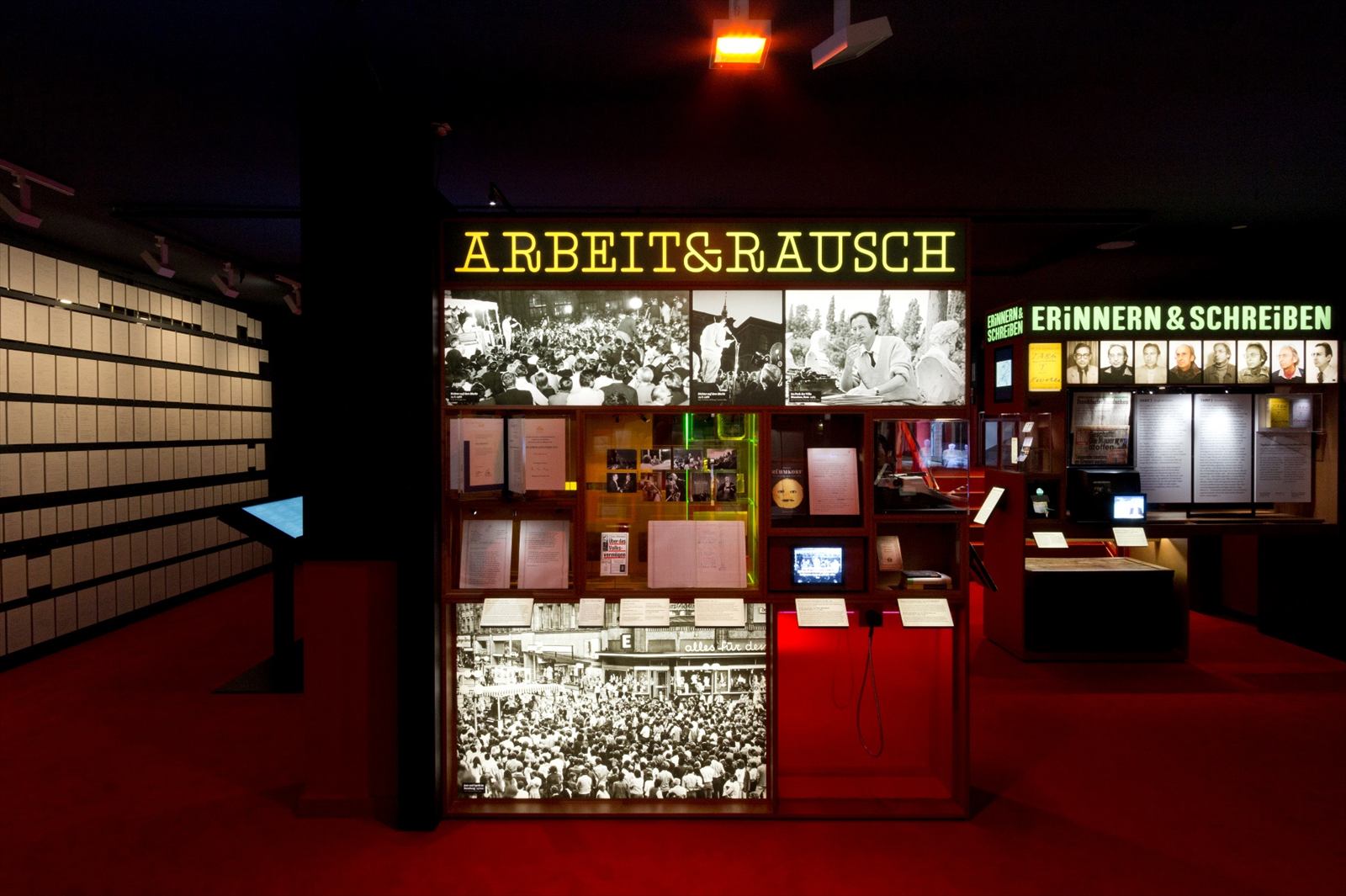 Themeninsel „Arbeit & Rausch“ mit Fotos, Vitrinen und kleinen Monitoren auf rotem Boden