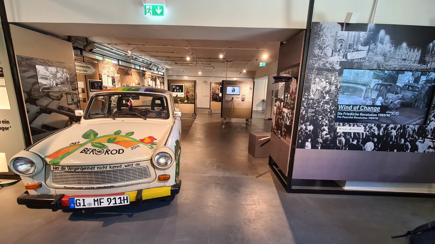 Ausstellungsraum mit bemaltem Trabant, dahinter Bildwände und Medienstationen
