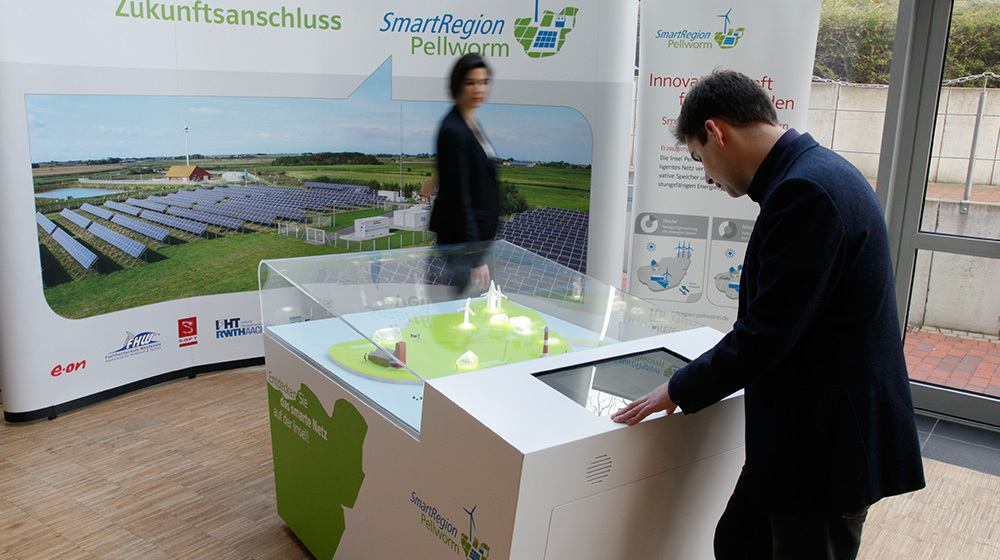 Besucher am Exponat mit beleuchtetem Inselmodell und Touchscreen vor einer Fotowand mit Solarpark