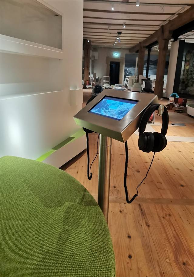 Touchscreen-Pult mit Kopfhörern im Ausstellungsraum mit Holzboden und Holzbalkendecke