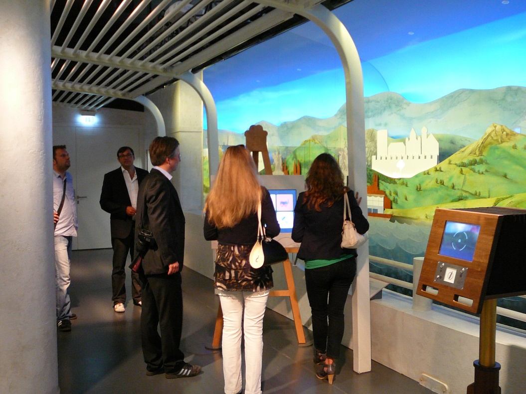 Besucher vor gemaltem Rheinpanorama mit Burg, rechts eine Medienstation im Holzgehäuse