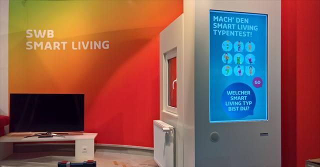 Raum mit Wandschriftzug „SWB Smart Living“, Fernseher und Typenquiz-Terminal mit Hochformat-Bildschirm