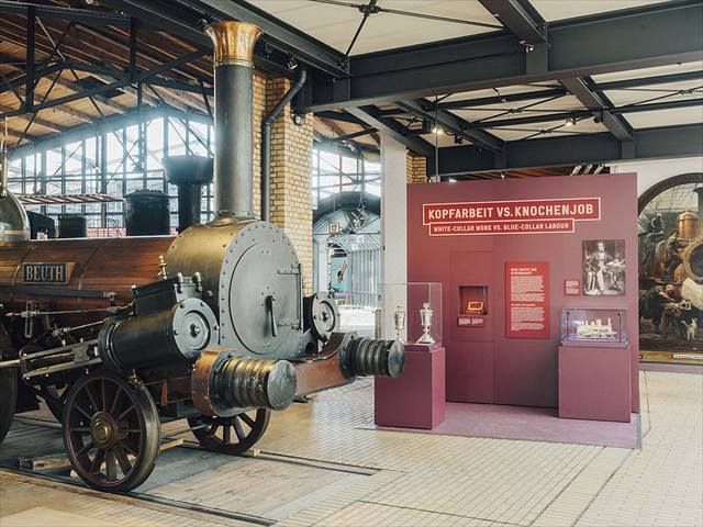 Historische Dampflokomotive in der Ausstellungshalle neben dem Themenbereich „Kopfarbeit vs. Knochenjob“