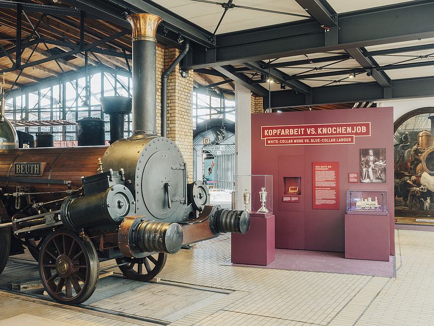 Historische Dampflokomotive in der Ausstellungshalle neben dem Themenbereich „Kopfarbeit vs. Knochenjob“