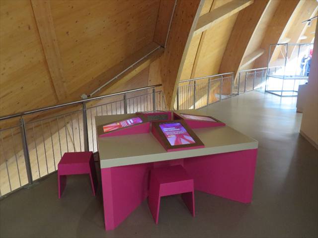 Weiß-pinkfarbener Tisch mit eingelassenen Touchscreens unter einer Holzdachkonstruktion