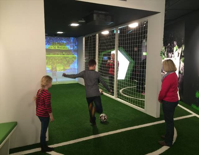 Kind schießt auf Kunstrasen einen Ball vor einer Projektionswand, rechts eine Netzwand