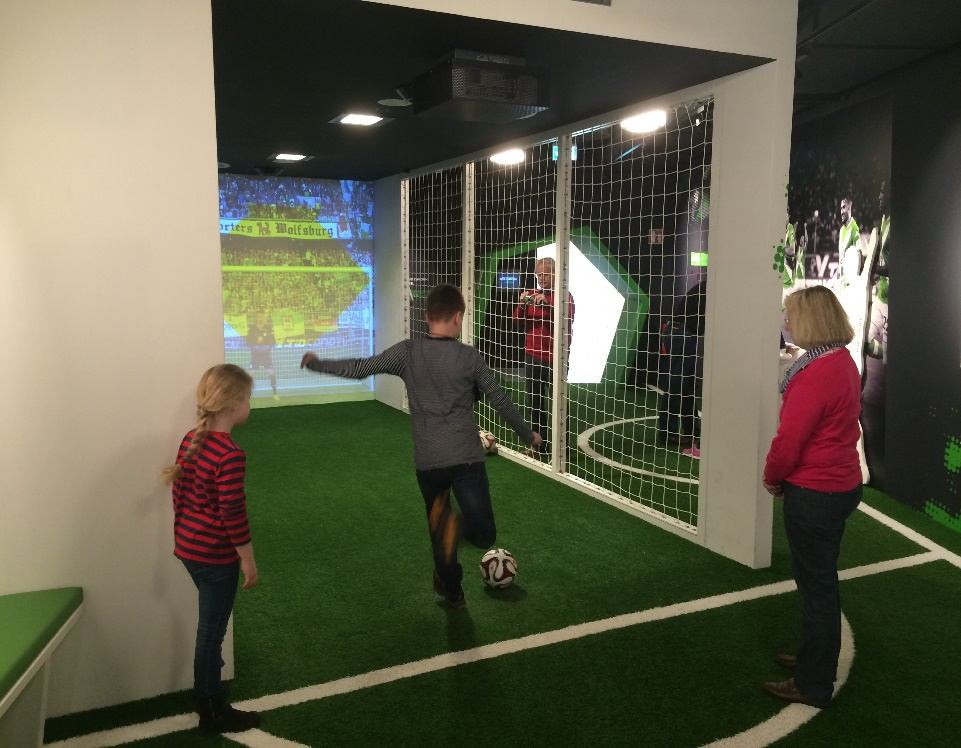 Kind schießt auf Kunstrasen einen Ball vor einer Projektionswand, rechts eine Netzwand